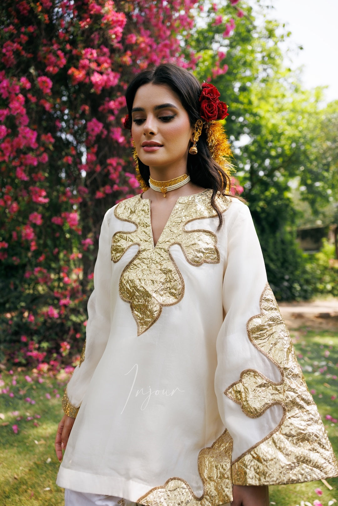 A woman wearing a cream-colored traditional kurta with ornate gold embroidery, standing outdoors in front of a lush garden with pink flowers.