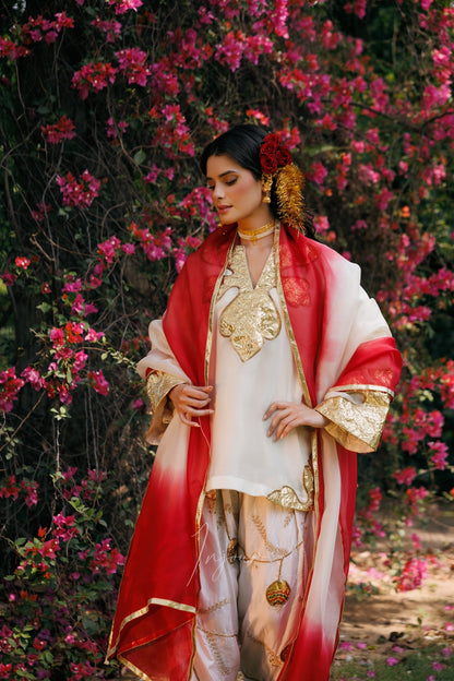 A woman wearing a traditional South Asian outfit with a red and white dupatta, gold-embroidered white kurta, and matching pants, standing in front of a dense background of pink flowers.