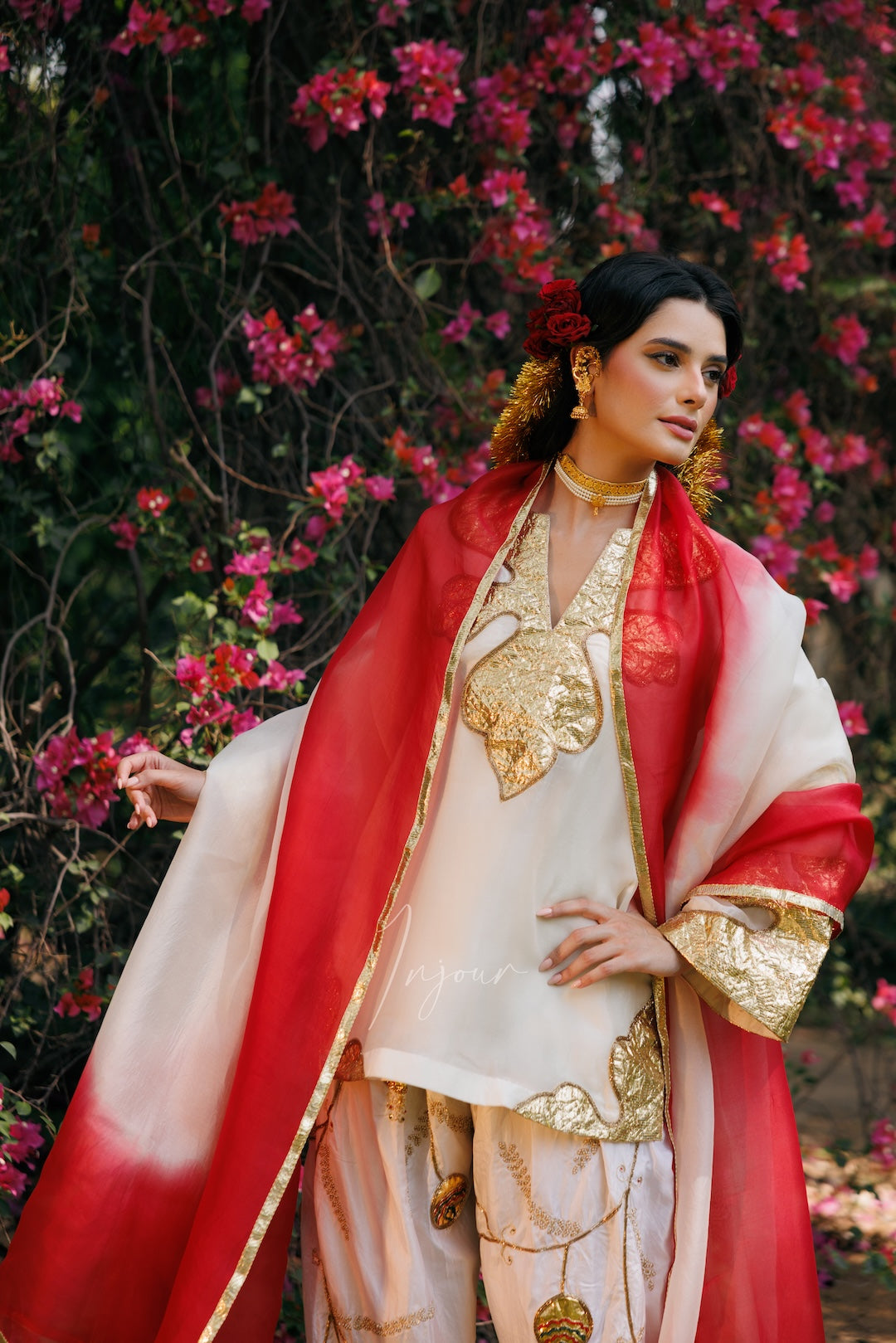 A woman wearing a traditional Indian outfit with a red and white dupatta, cream-colored kurta with gold embroidery, and patterned pants, standing in front of a background of pink flowers.