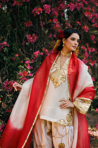 A woman wearing a traditional Indian outfit with a red and white dupatta, cream-colored kurta with gold embroidery, and patterned pants, standing in front of a background of pink flowers.