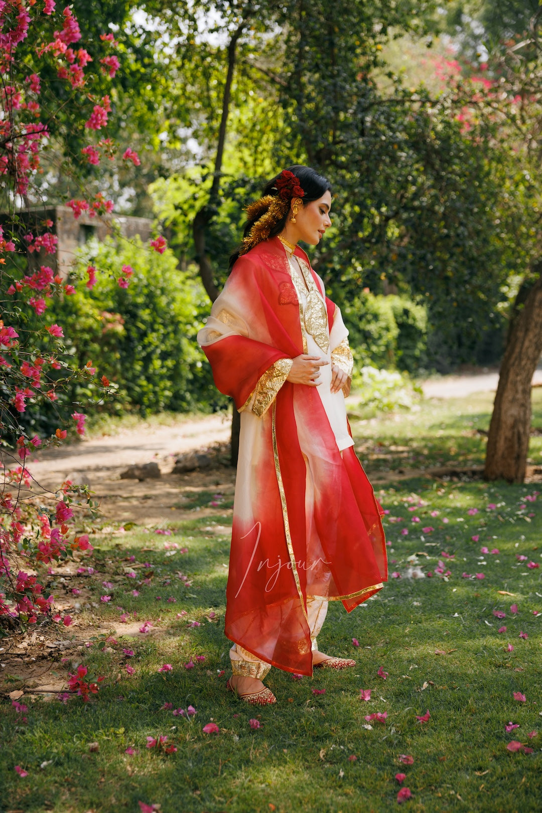 A woman wearing a traditional red and white embroidered kurta with a matching dupatta stands in a garden setting with green grass, trees, and pink flowers.