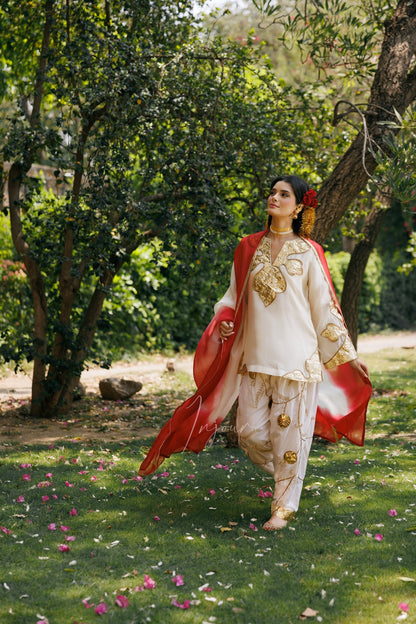 A woman wearing a traditional white embroidered outfit with gold detailing and a flowing red dupatta stands outdoors in a garden with trees and grass.