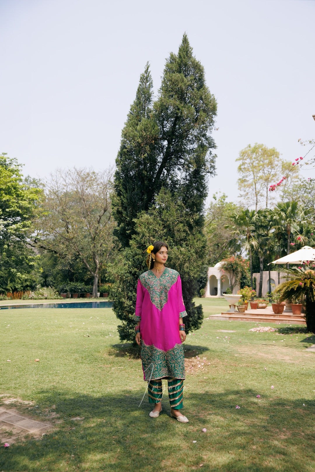 A woman stands outdoors in a large garden wearing a bright pink kurta with green printed embroidery, paired with green patterned pants. The background features a manicured lawn, a tall central tree, and a pool in the distance.