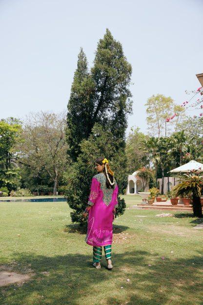 A woman stands on a grassy lawn wearing a bright pink traditional outfit with green striped pants, photographed outdoors in a garden setting with trees and a pool in the background.