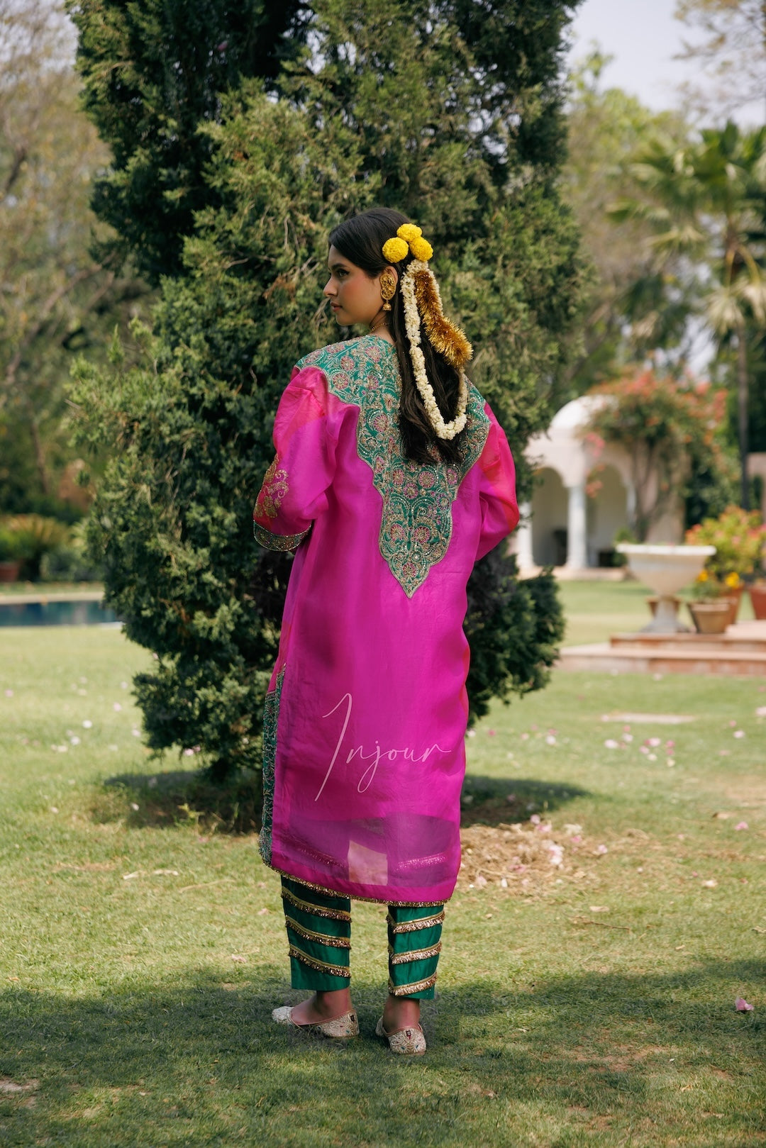 A woman stands outdoors in a garden wearing a bright pink traditional outfit with green and gold embroidered details on the back, paired with green striped pants. She has yellow flowers in her hair.