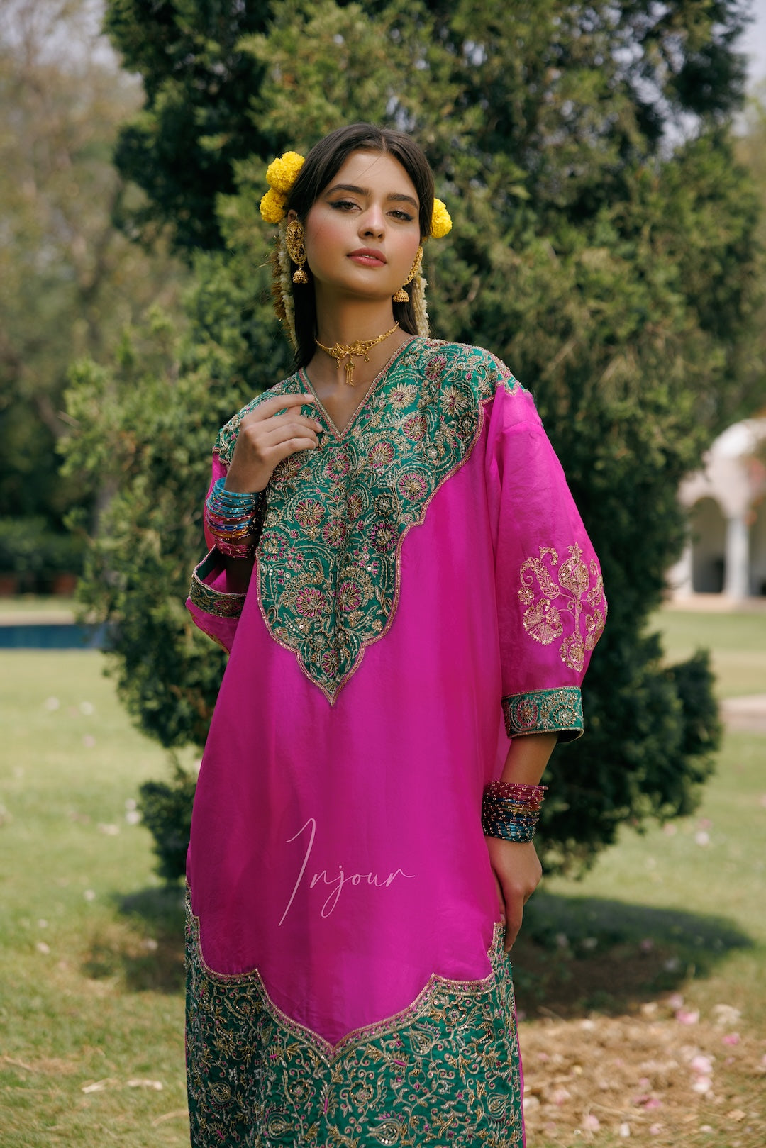 A woman wearing a bright pink and green embroidered traditional kurta with floral patterns, standing outdoors in a park with trees and grass in the background.
