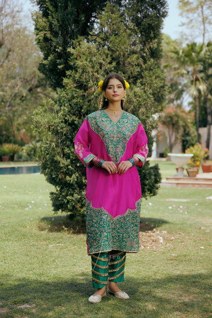 A woman stands outdoors in a garden wearing a bright pink and green traditional outfit with ornate embroidery, striped green pants, and yellow flower hair accessories.
