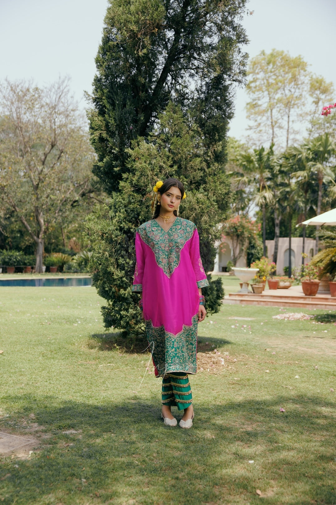 A woman stands in a garden wearing a bright pink traditional kurta with green embroidered detailing, paired with green and black striped pants.
