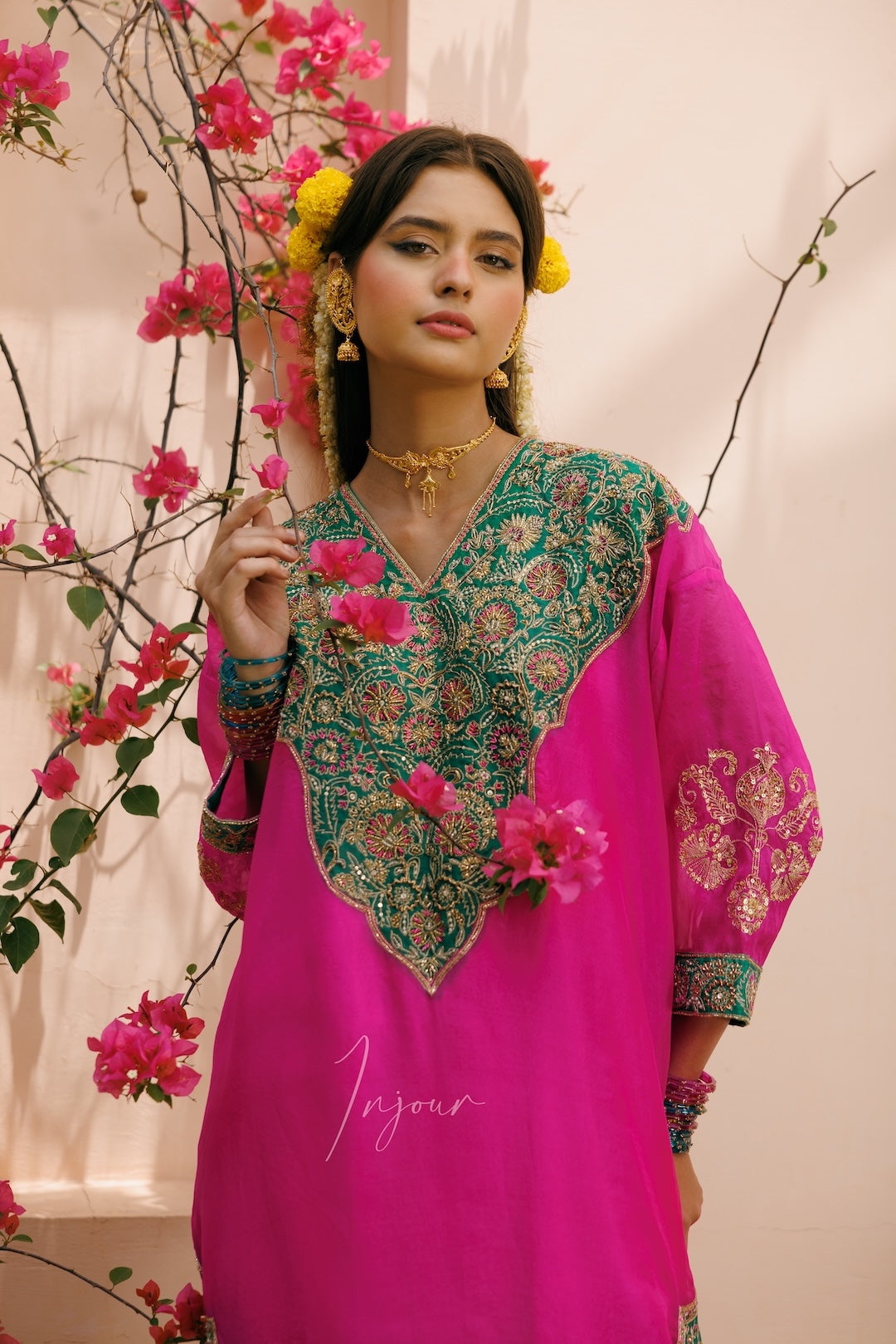A woman modeling a bright pink traditional outfit with an ornate green and gold embroidered yoke, standing against a light beige wall decorated with pink flowers.
