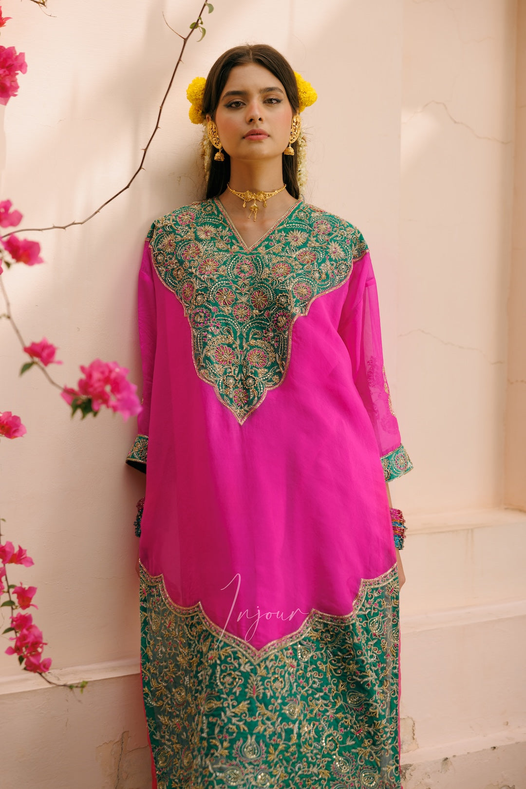 A woman wearing a bright pink traditional kurta with ornate green and gold embroidery, standing against a light beige wall with pink flowers on the left side.