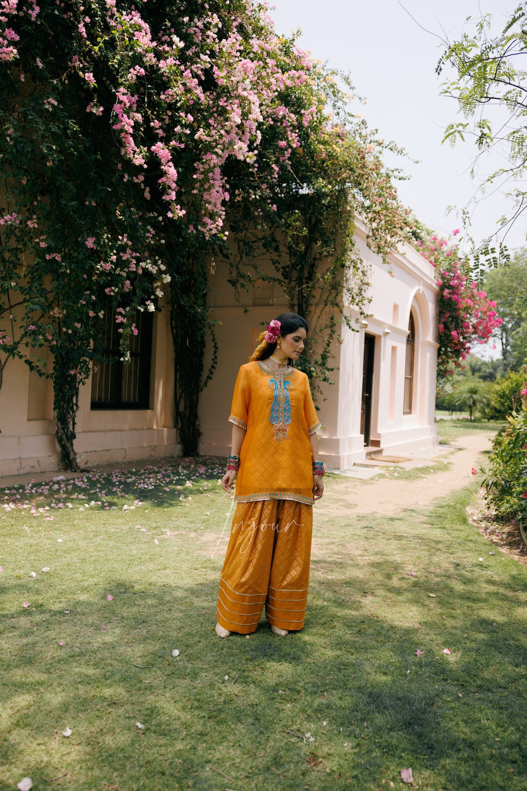A woman stands on a grassy lawn in front of a beige building covered with pink flowers, wearing a bright mustard yellow traditional outfit with a blue embroidered design on the chest.