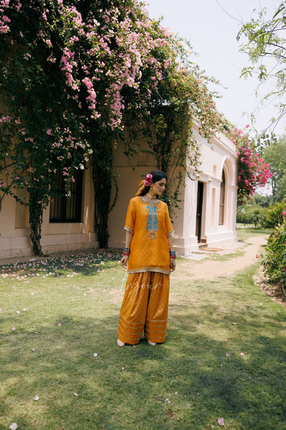 A woman stands on a grassy lawn in front of a beige building covered with pink flowers, wearing a bright mustard yellow traditional outfit with a blue embroidered design on the chest.