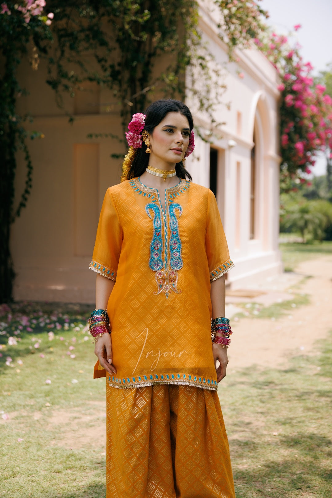 A woman stands outdoors in front of a light-colored building, wearing a bright mustard yellow traditional outfit with blue and silver embroidery on the chest, matching wide-leg pants, and colorful jewelry.