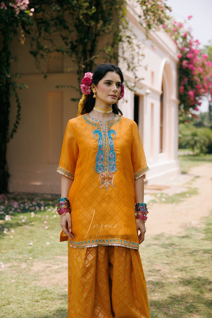 A woman stands outdoors in front of a light-colored building, wearing a bright mustard yellow traditional outfit with blue and silver embroidery on the chest, matching wide-leg pants, and colorful jewelry.