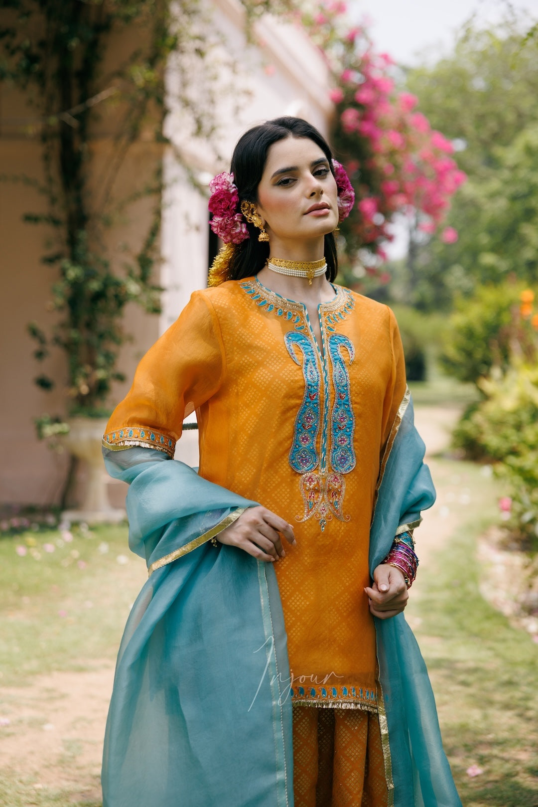 A woman wearing a traditional South Asian outfit with a mustard yellow embroidered kurta, a turquoise dupatta, and matching trousers, standing in a garden setting.