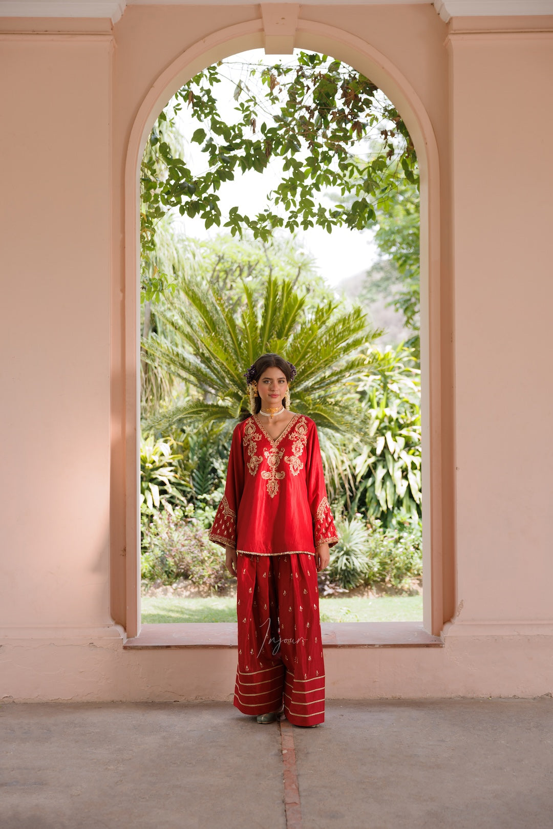 A woman stands in a peach-colored arched corridor wearing a traditional red embroidered kurta and wide-leg pants set, with a lush green garden visible through the opening behind her.