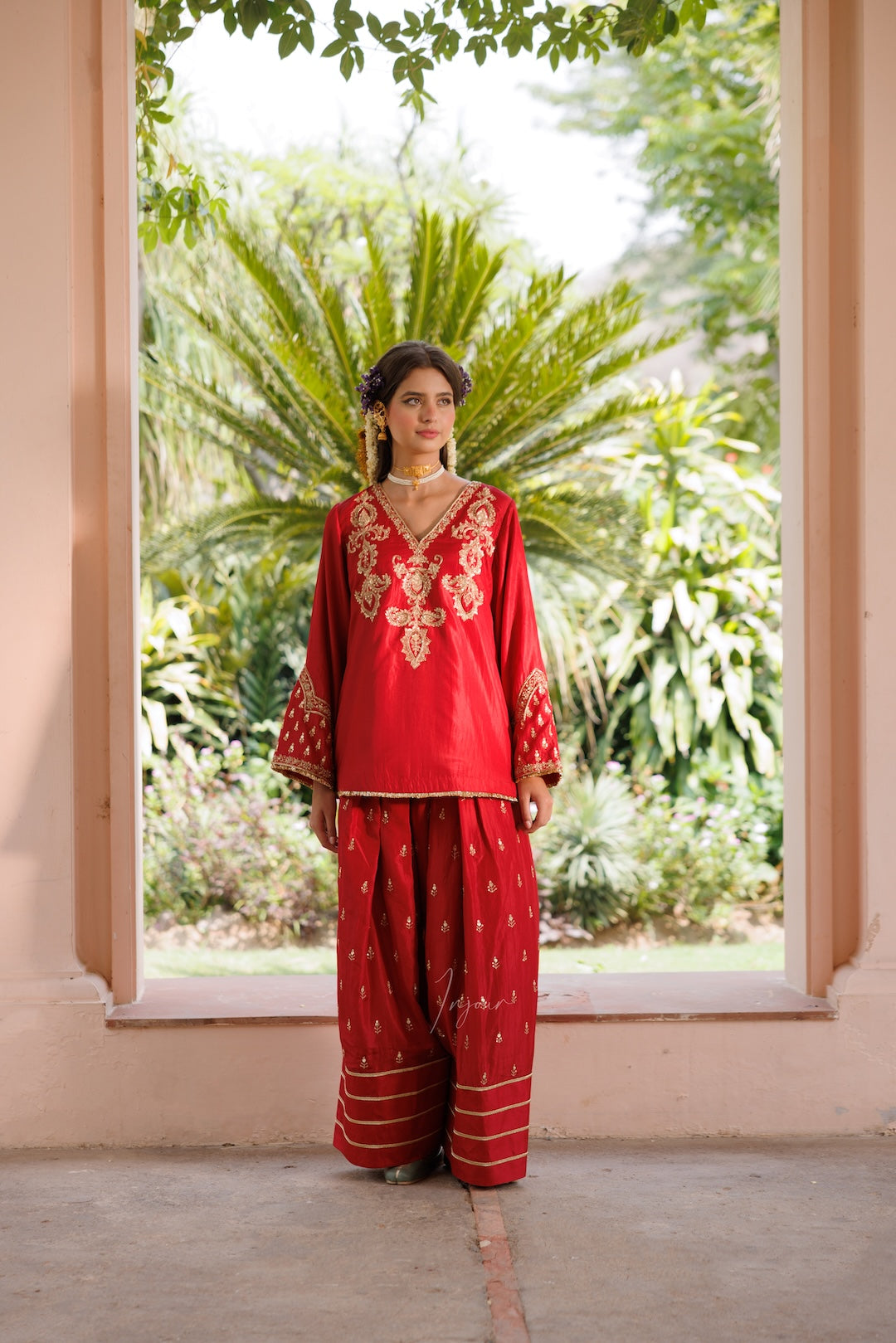 A woman stands in a doorway wearing a red traditional outfit with gold embroidery and wide-leg pants, with a garden visible behind her.