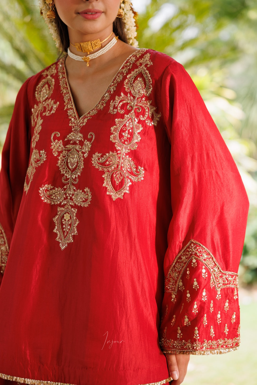 A close-up of a woman wearing a bright red traditional tunic with ornate gold embroidery around the neckline, sleeves, and cuffs, photographed outdoors against a blurred green background.