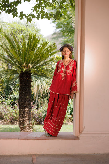 A woman wearing a red traditional embroidered outfit with gold detailing stands in a doorway, with a lush green garden and palm tree visible behind her.