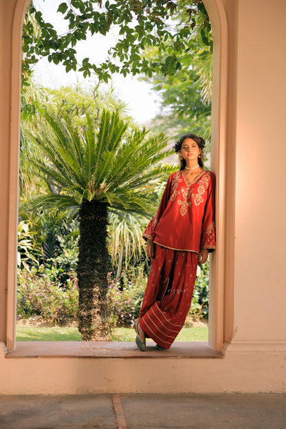 A woman wearing a red embroidered traditional outfit with wide sleeves and loose pants, standing in a doorway with a lush garden and palm tree visible behind her.