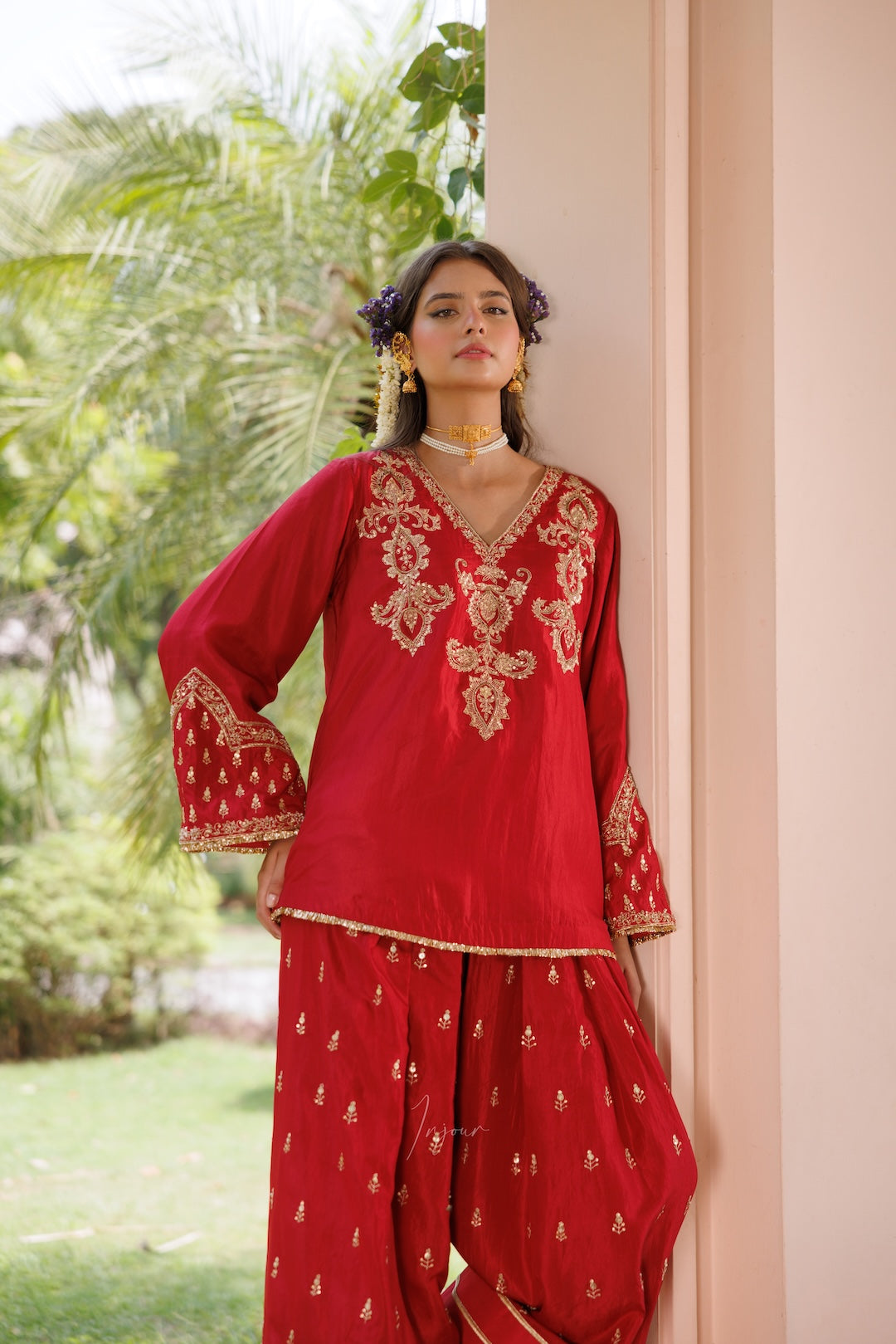 A woman models a red traditional Indian outfit with gold embroidery, featuring a V-neck kurta with ornate embroidery on the chest and sleeves, paired with matching red pants, set against a background of a beige wall and green foliage.