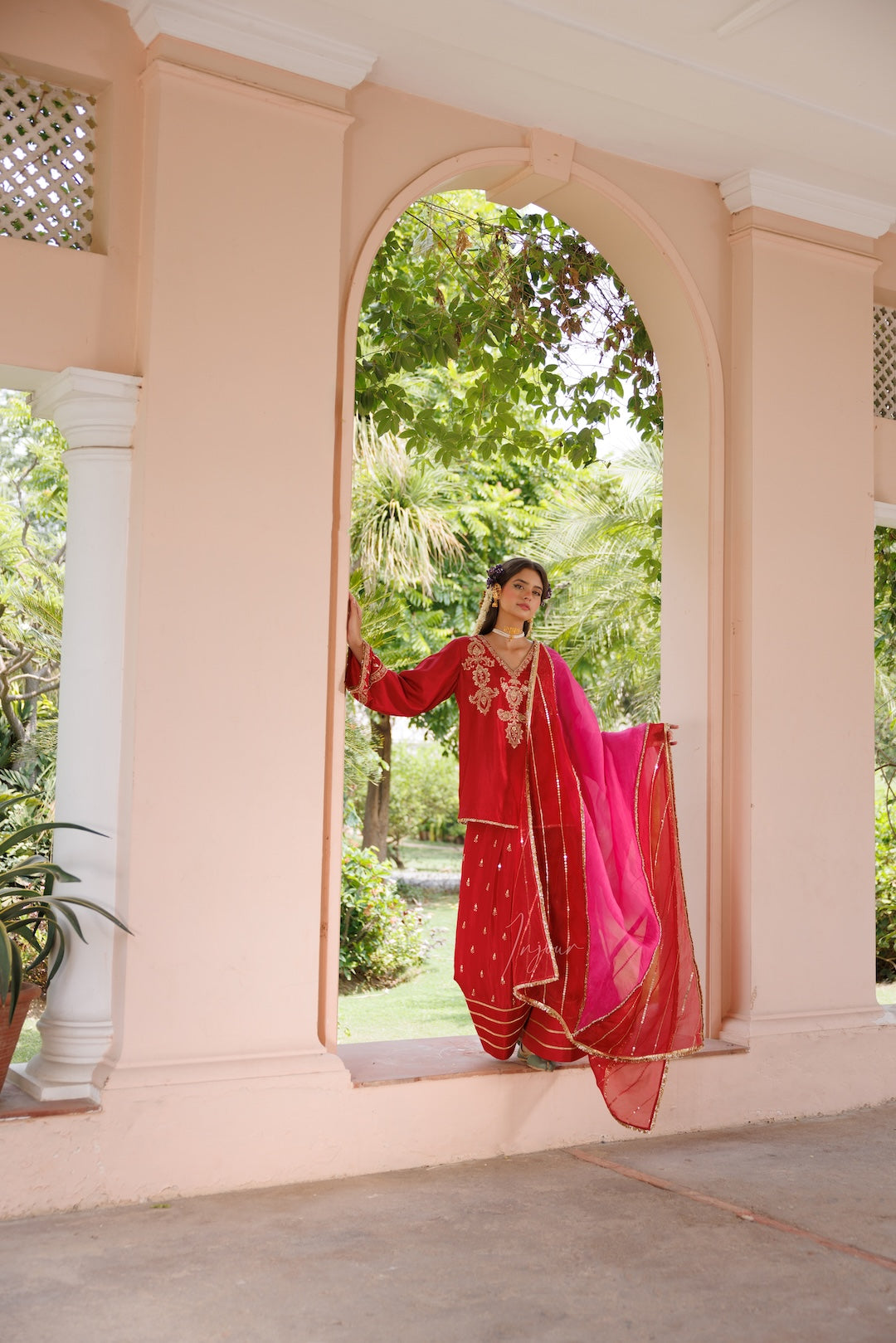 A woman wearing a traditional red and pink embroidered salwar kameez outfit with a bright pink dupatta stands in an arched doorway of a peach-colored building, with green garden foliage visible behind her.