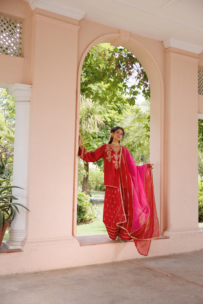 A woman wearing a traditional red and pink embroidered salwar kameez outfit with a bright pink dupatta stands in an arched doorway of a peach-colored building, with green garden foliage visible behind her.