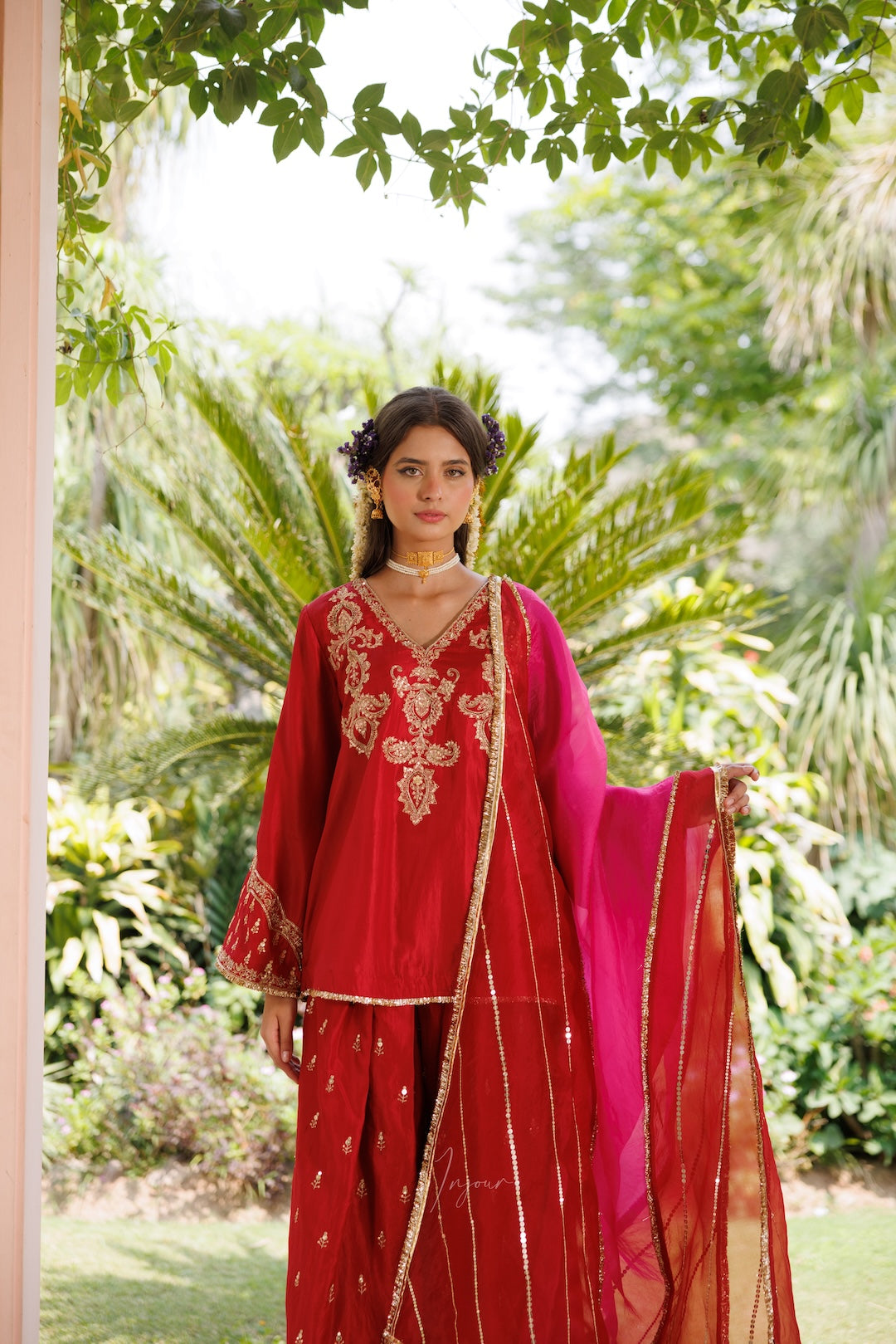 A woman stands outdoors in a lush garden wearing a traditional red ethnic outfit with gold embroidery, a bright pink dupatta, and matching red pants. She is framed by green leaves hanging from above.