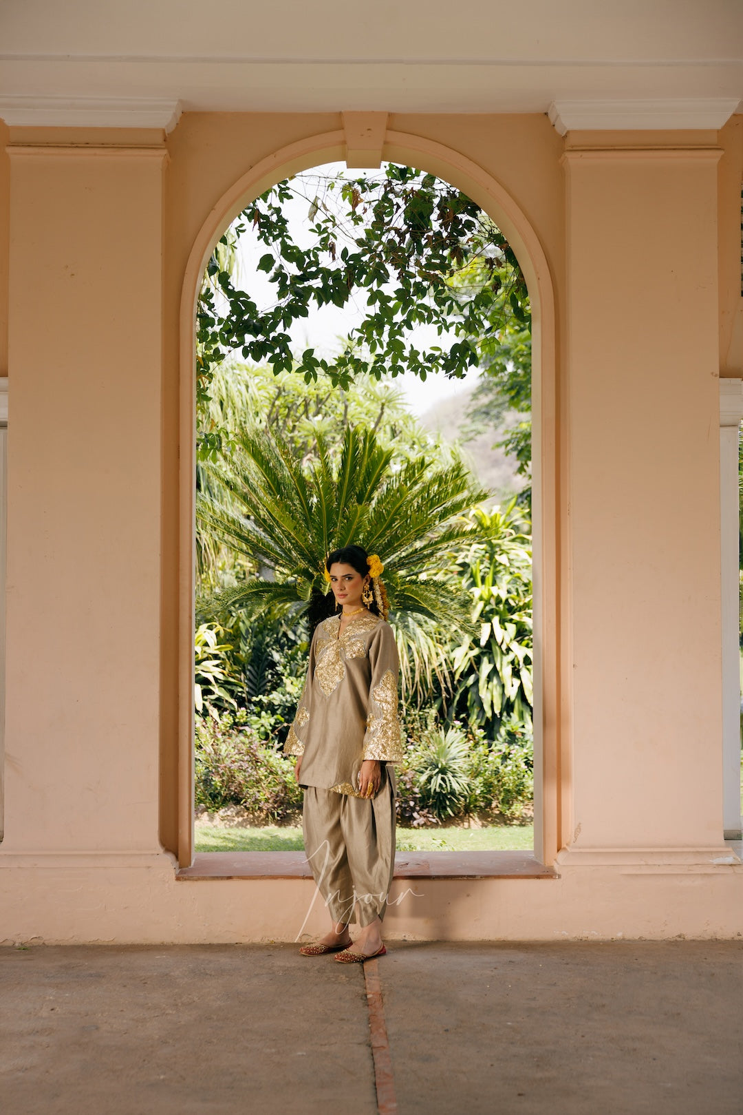 A woman wearing a beige traditional outfit with gold embroidery stands in a doorway, looking back over her shoulder. The background features a lush green garden visible through an arched opening, framed by peach-colored walls.