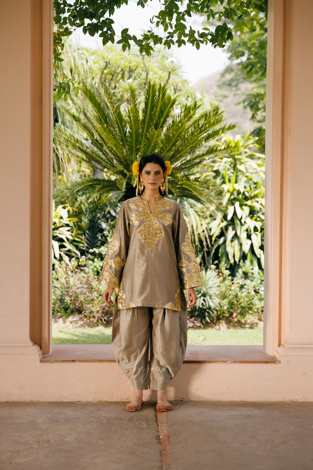 A woman stands barefoot in a doorway wearing a beige and gold embroidered traditional outfit, with lush green tropical plants visible in the background.