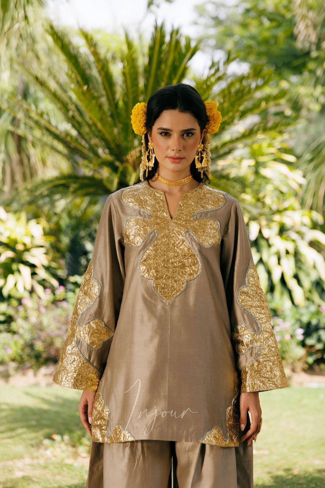 A woman stands outdoors in a lush garden wearing a taupe-beige traditional outfit with ornate gold embroidery and matching gold accessories.