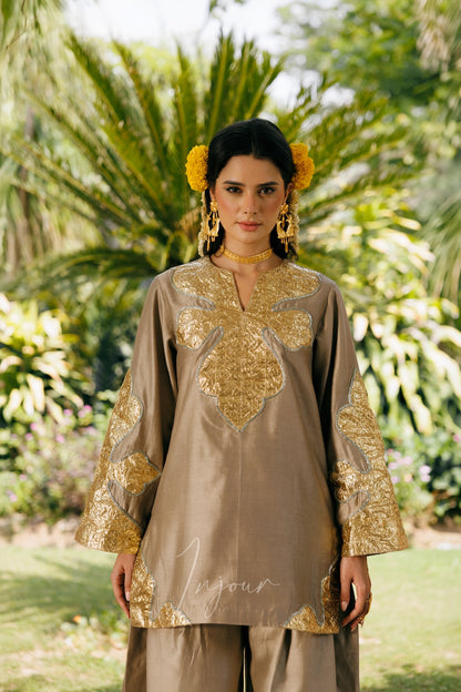 A woman stands outdoors in a lush garden wearing a taupe-beige traditional outfit with ornate gold embroidery and matching gold accessories.