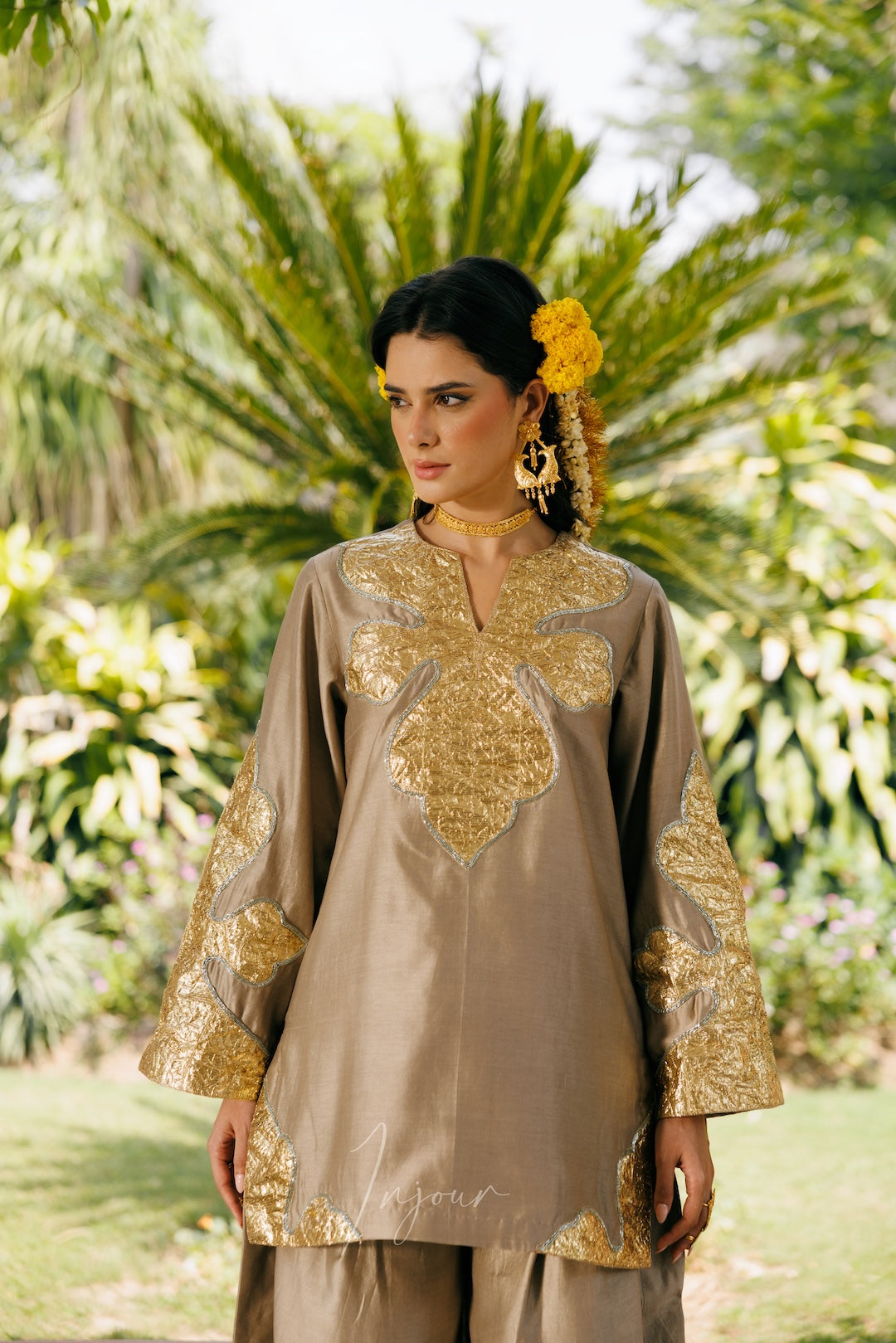 A woman wearing a traditional beige tunic with ornate gold embroidery and matching pants, standing outdoors in front of a lush green tropical background.