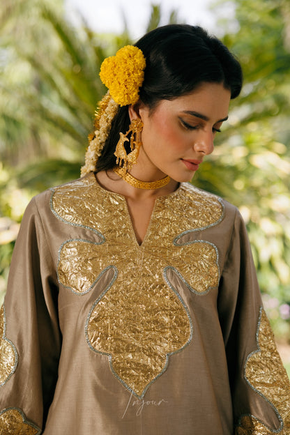A woman wearing a taupe-brown traditional tunic with large metallic gold embroidered motifs and a yellow flower in her hair, photographed outdoors against a soft green foliage background.