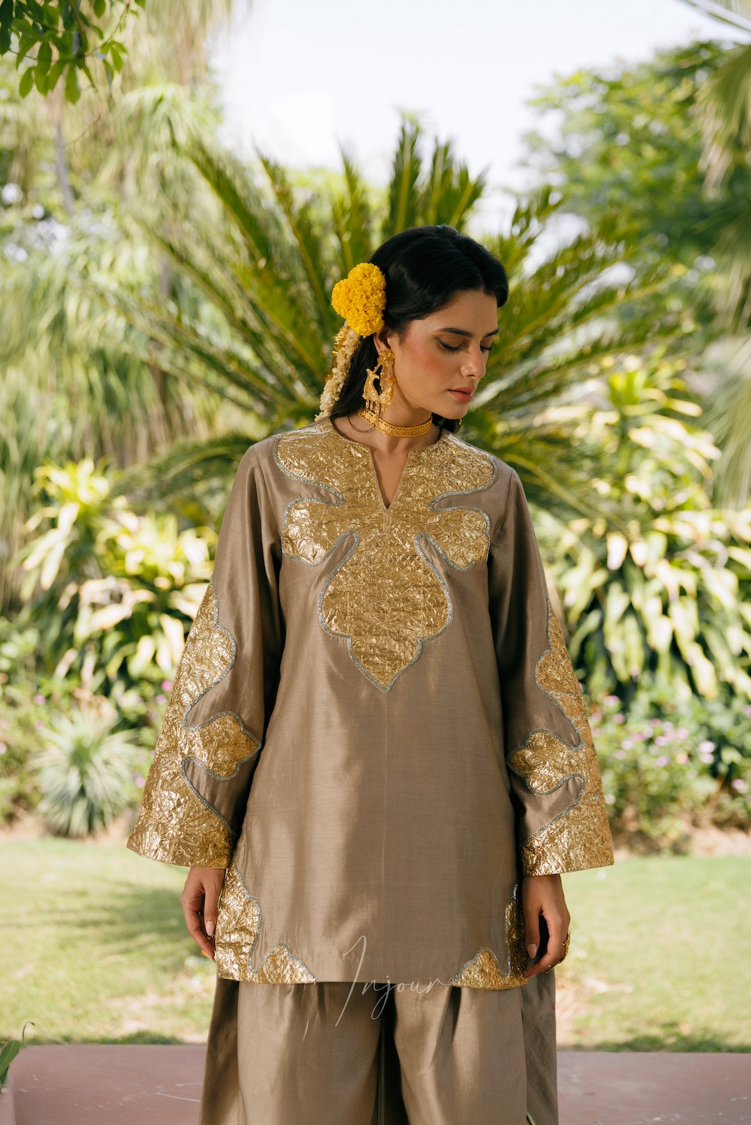 A woman wearing a brown traditional kurta set with ornate gold embroidery, standing outdoors in front of a lush green garden background.