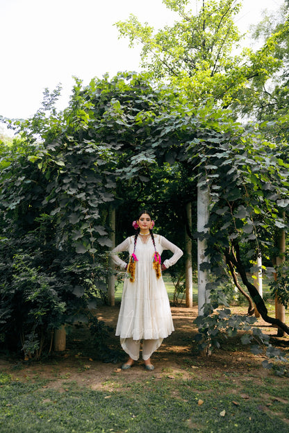 A woman stands outdoors under a leafy vine archway, wearing a long white traditional dress with pink and orange floral garlands around her neck.