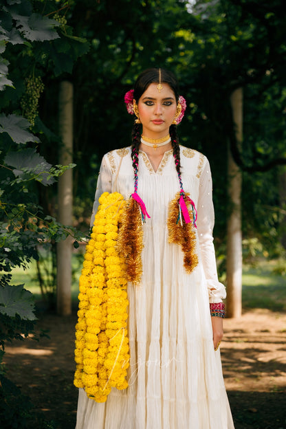 A woman stands outdoors in a lush green setting wearing a traditional white embroidered dress and holding a large yellow marigold garland. She has pink flowers in her hair and brown tinsel braid accessories.