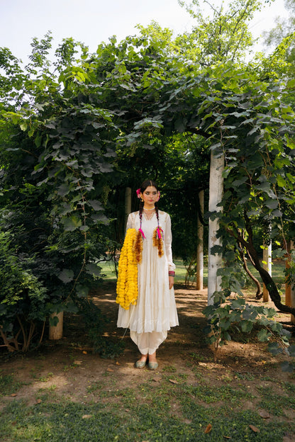 A woman stands outdoors under a leafy vine archway wearing a white traditional outfit with a bright yellow floral garland draped over her shoulder and pink flower accents in her hair.