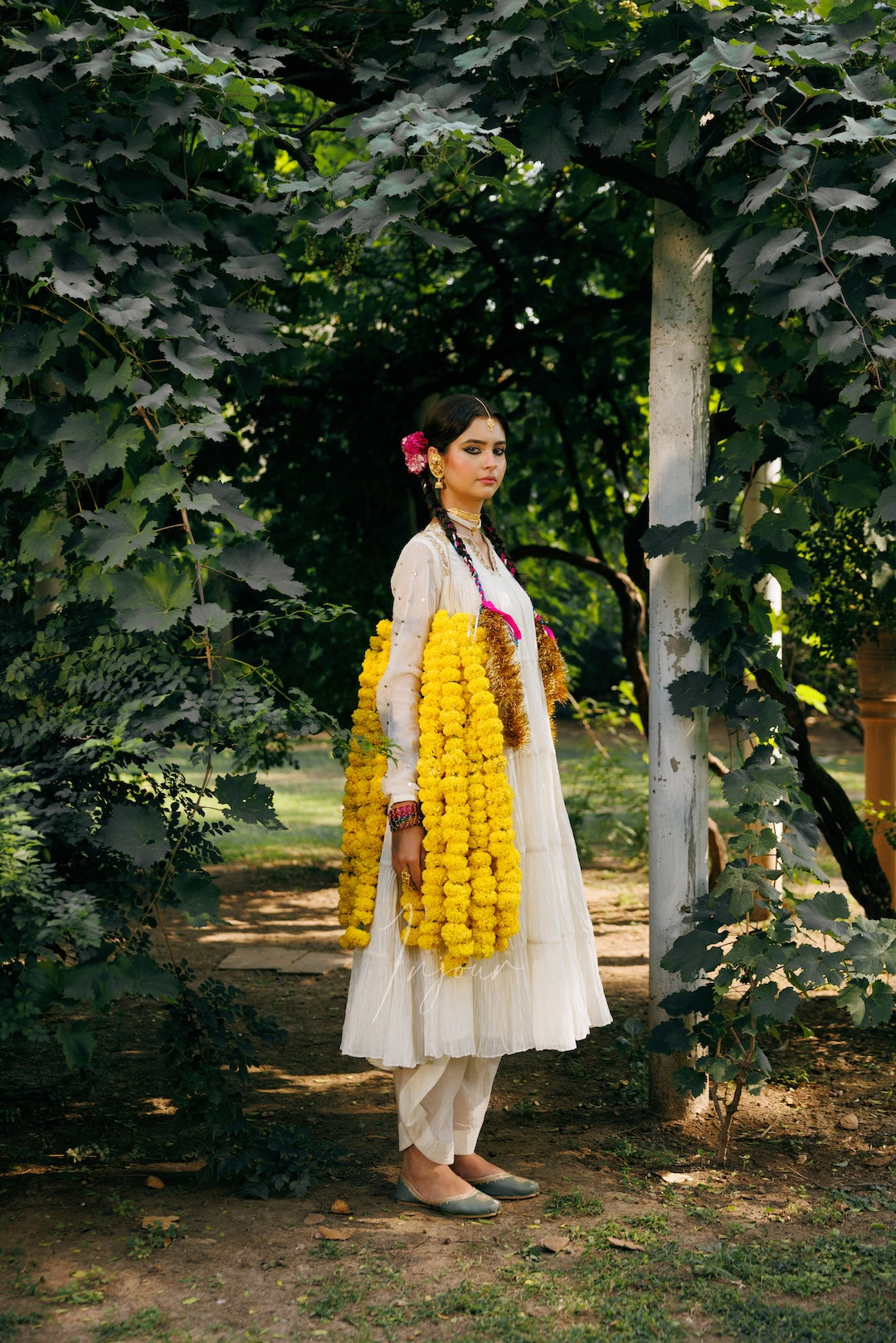 A woman stands outdoors in a garden wearing a white traditional outfit with a long yellow flower garland draped over her shoulders and a pink flower in her hair.