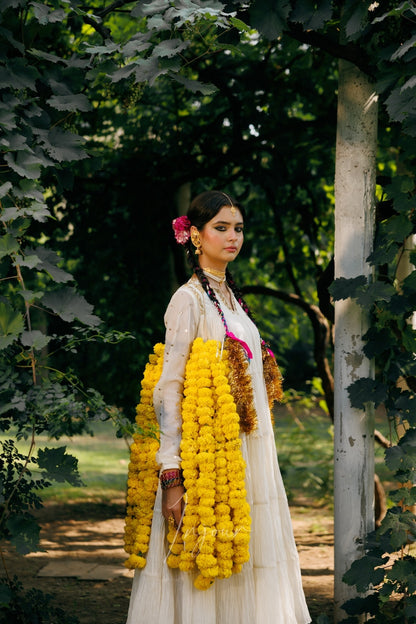A woman standing outdoors in a garden or forest setting, wearing a long white traditional dress and holding large yellow flower garlands. Trees and greenery fill the background.