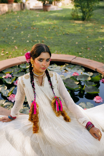 A woman wearing a traditional white embroidered dress with pink and gold tassel hair accessories sits by a pond with lily pads and pink flowers in an outdoor garden setting.