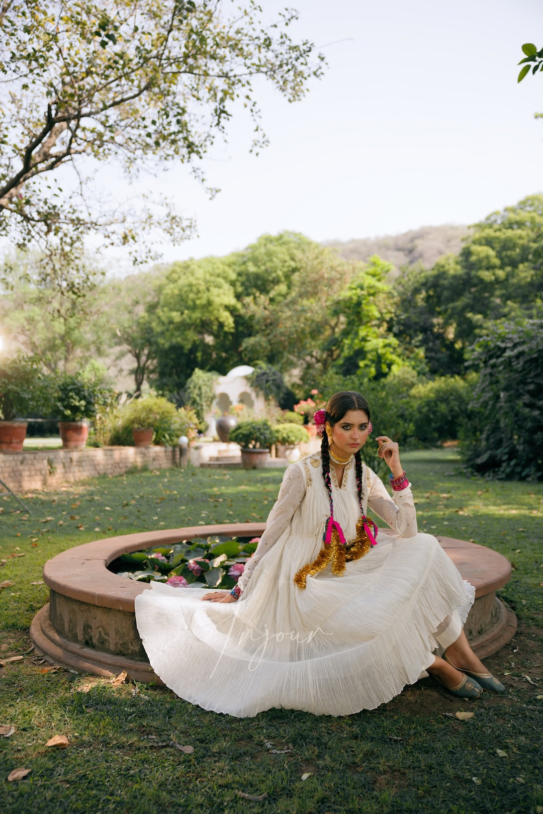 A woman in a flowing white traditional dress sits on a stone planter in a lush green outdoor garden setting.