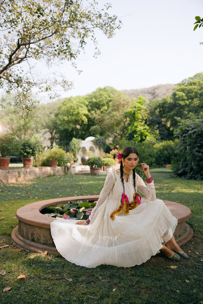 A woman in a flowing white traditional dress sits on a stone planter in a lush green outdoor garden setting.