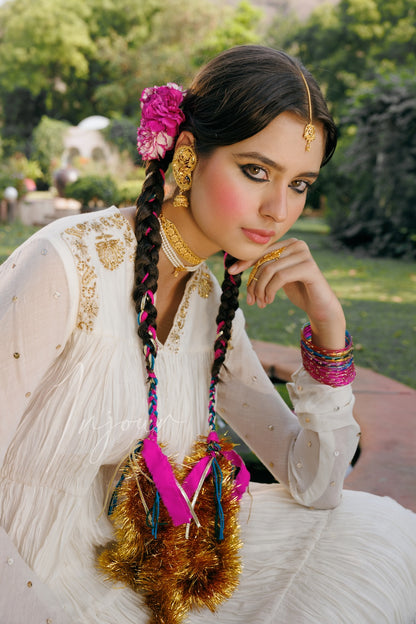 A woman wearing traditional Indian attire, featuring a white embroidered kurta, gold jewelry, and a bright pink flower in her braid, posed outdoors in a garden setting.
