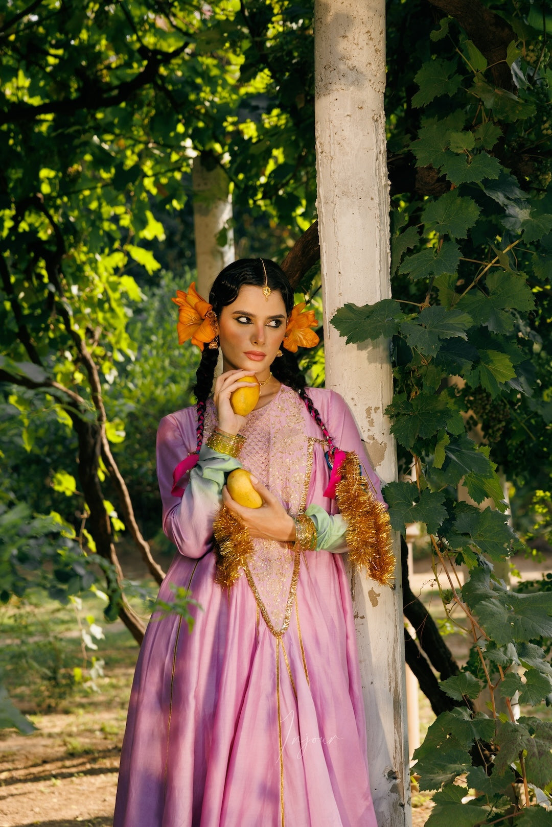 A woman in a lavender and pink traditional dress with orange flowers in her hair, holding yellow fruits, posing outdoors next to a tree trunk in a lush green forest.