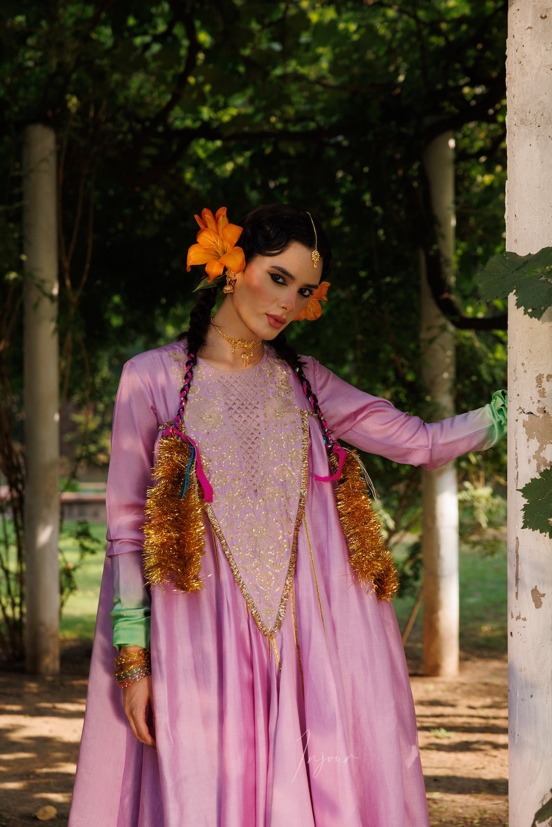 A woman wearing a traditional pink and purple embroidered dress with gold tassel accents, standing outdoors in a garden setting.