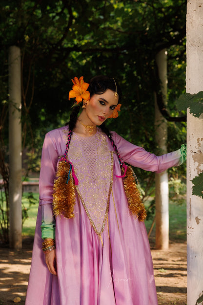 A woman wearing a traditional pink and purple embroidered dress with gold tassel accents, standing outdoors in a garden setting.