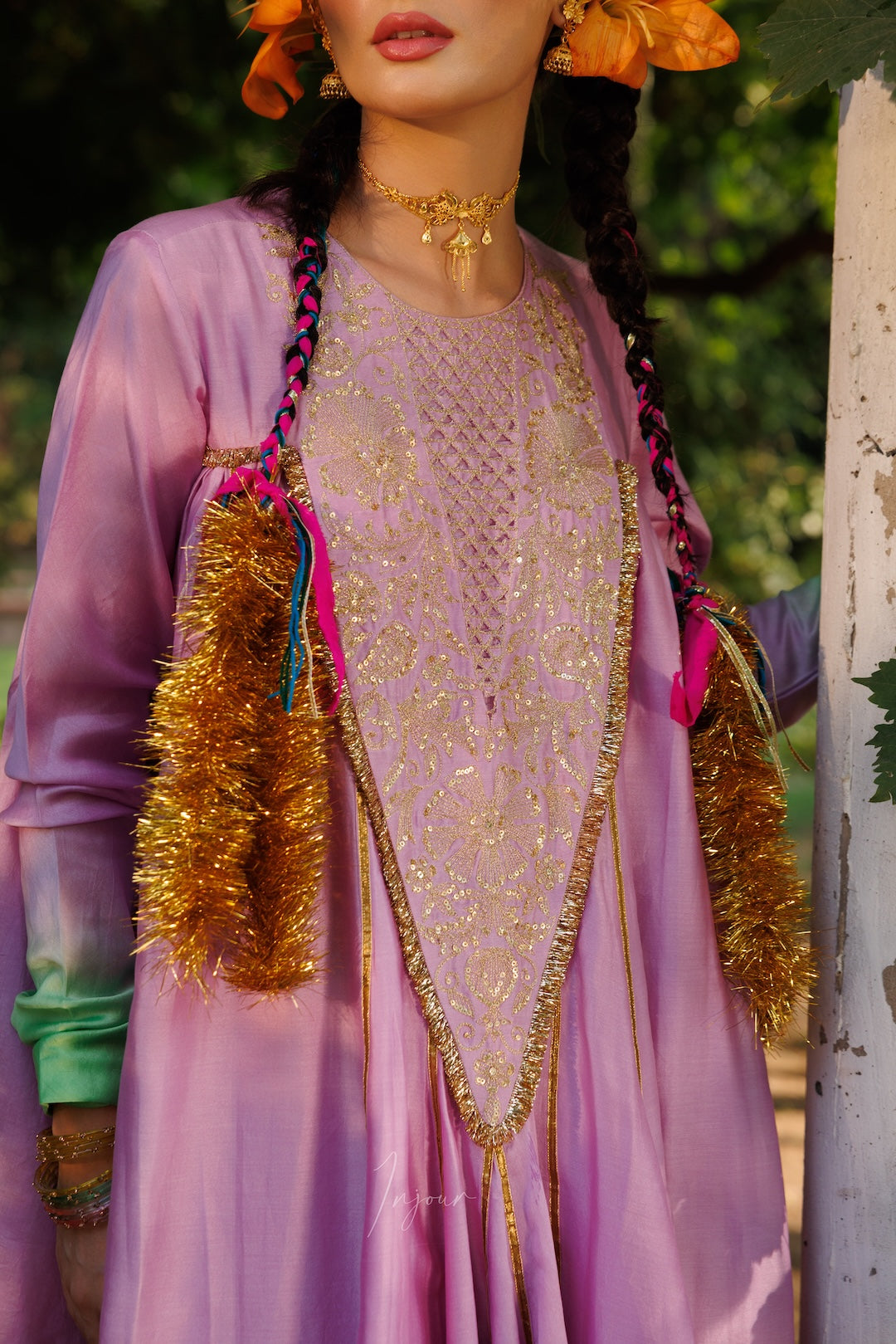 A close-up of a model wearing a traditional South Asian outfit featuring a lavender purple kurta with intricate gold embroidery, a sheer beige mesh inner layer, and decorative gold tinsel tassels with pink and teal ribbons.