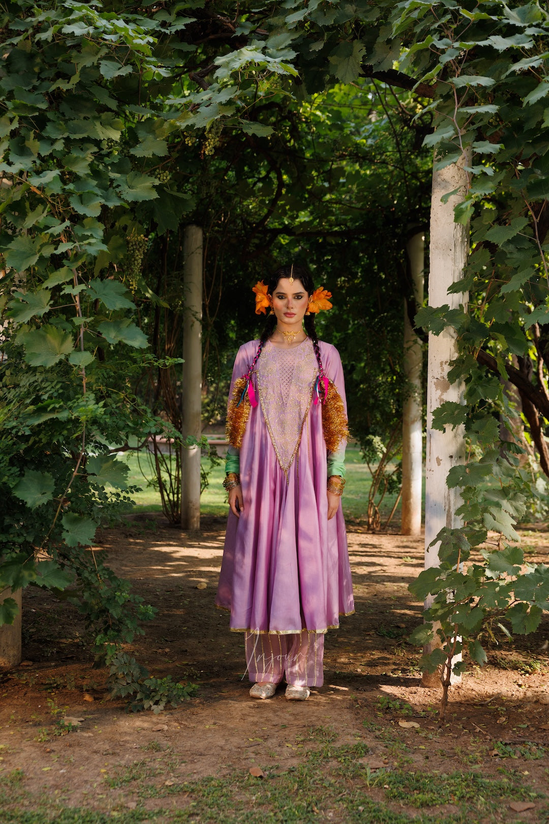 A woman stands in a garden pathway wearing a lavender traditional outfit with orange and gold embroidered details, green sleeves, and orange floral hair ornaments.
