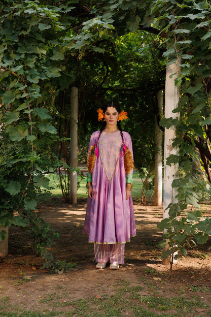 A woman stands in a garden pathway wearing a lavender traditional outfit with orange and gold embroidered details, green sleeves, and orange floral hair ornaments.